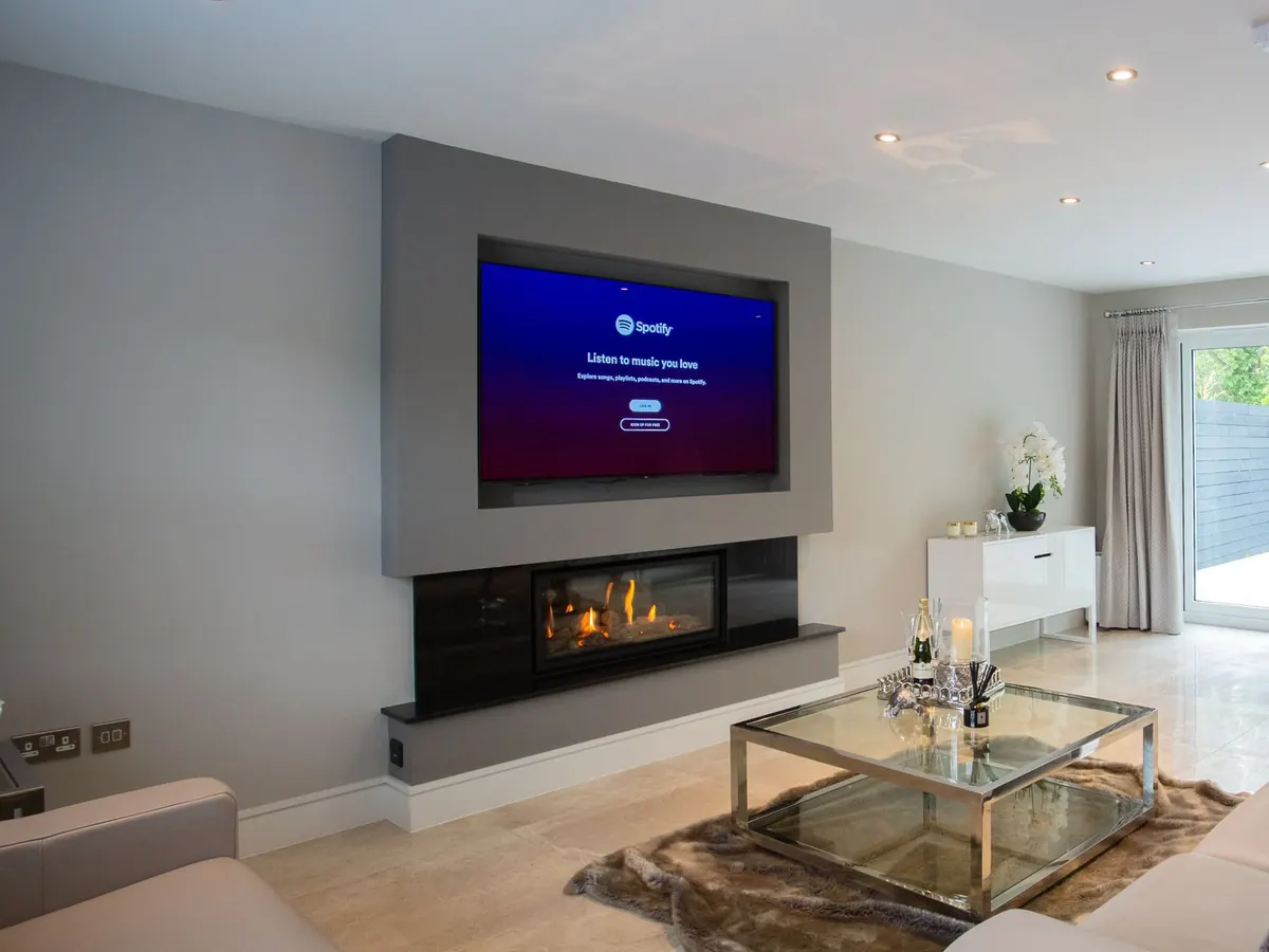 TV mounted on a grey panel above a linear fireplace in a bright living room with a glass coffee table