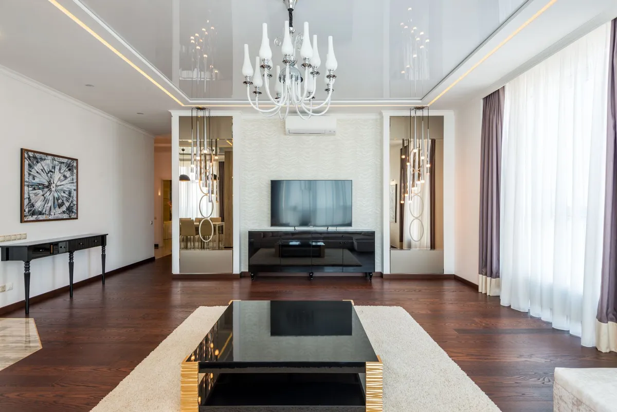 TV on a black console between two mirrored niches, under a white chandelier, in a living room with dark wood floors