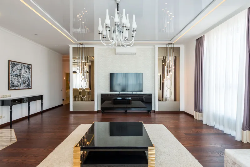 TV on a black console between two mirrored niches, under a white chandelier, in a living room with dark wood floors