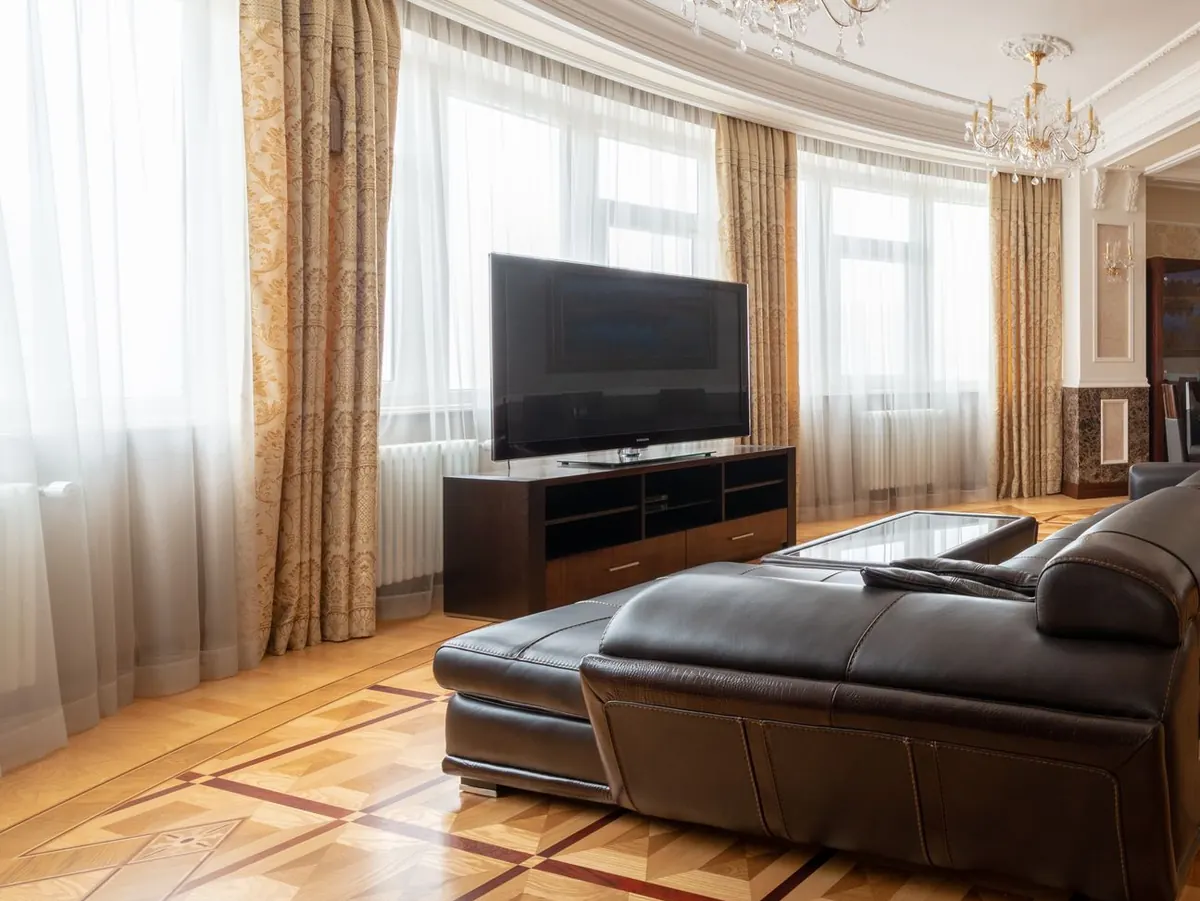 TV on a low stand in a large living room with ornate plaster ceilings, parquet floors and a brown leather sectional